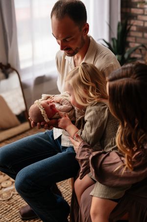 A father holds a newborn baby while a young girl and mother lean in to admire the infant in a cozy living room.