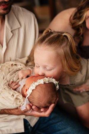 A young girl with Down syndrome lovingly kisses a newborn baby wearing a floral headband, held by parents.