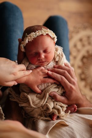 Newborn baby girl in floral headband and lace wrap, cradled gently by parents hands in a warm, cozy setting.