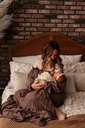A mother in a brown dress sits on a bed cradling her newborn baby against a rustic brick wall backdrop.