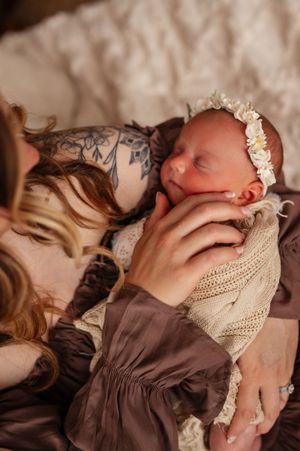 A tattooed mother tenderly holds her sleeping newborn wrapped in a knit blanket with a floral headband.