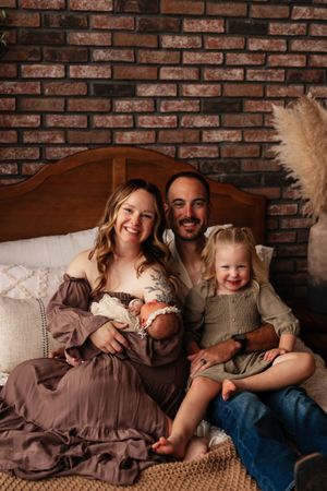 A smiling family of four poses on a bed with brick wall backdrop, mom in mauve dress holding newborn, dad with toddler girl.