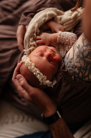 Newborn baby with floral headband cradled in parents hands, with tattoo visible, soft warm tones.