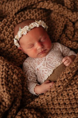 Sleeping newborn baby girl in white lace outfit and floral headband on brown chunky knit blanket.