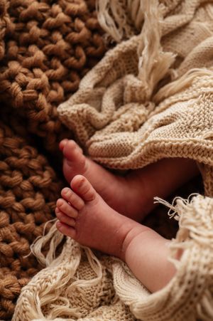Tiny newborn baby feet peeking out from cozy brown and cream knitted blankets in a warm, soft photography setup.