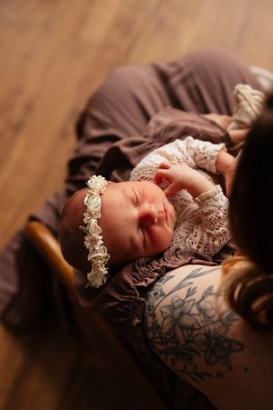Sleeping newborn baby girl wearing a white floral headband, cradled in a mothers tattooed arms on a mauve blanket.