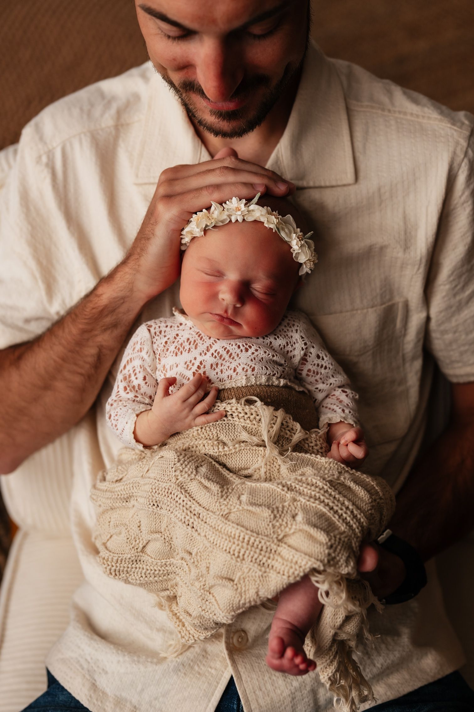 A smiling father tenderly holds his sleeping newborn baby girl dressed in a white lace outfit and floral headband.