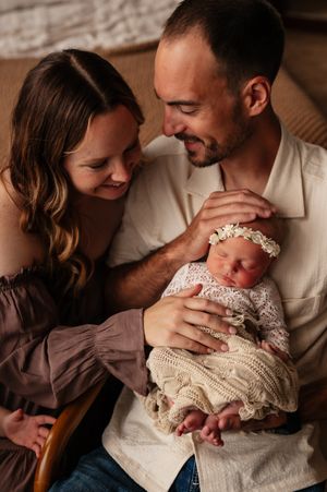 New parents lovingly gaze at their sleeping newborn baby girl in a lace outfit and floral headband.