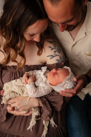 A couple admires their sleeping newborn dressed in lace, with the father's floral tattoo visible.