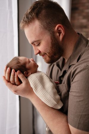 A bearded father tenderly holds his sleeping newborn baby near a window, smiling softly in a warm, intimate moment.