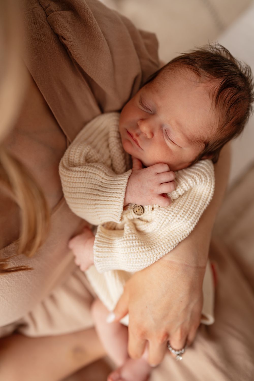 Sleeping newborn baby in cream knit outfit cradled in mother's arms during professional newborn photography session.