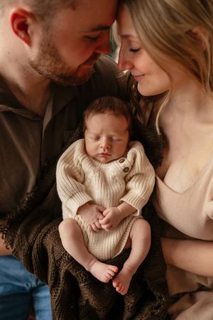 New parents tenderly touch foreheads while cradling their sleeping newborn dressed in cream knit romper.