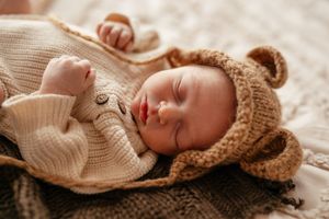 Peaceful newborn baby sleeping in a bear ear knit hat and cream sweater onesie on a cozy blanket.