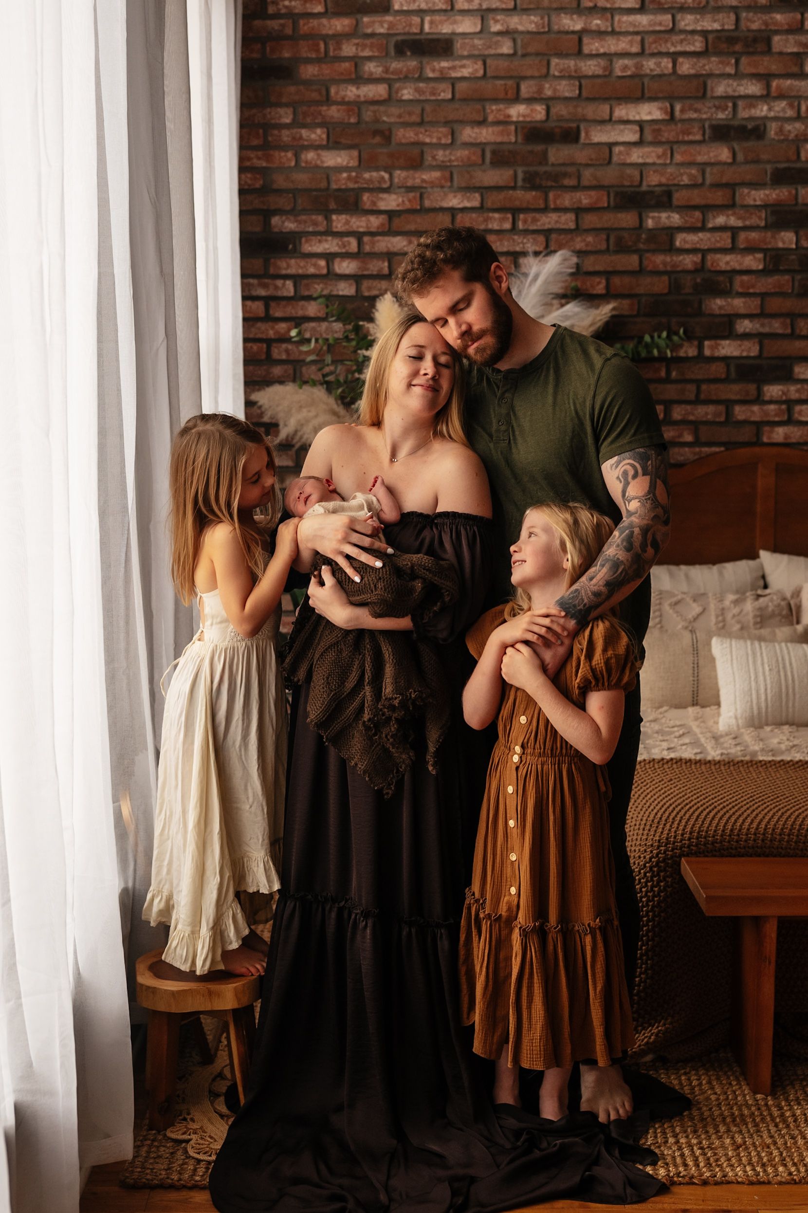 Family portrait with parents holding newborn, two daughters in dresses, brick wall background in cozy bedroom setting.