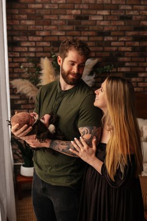 A couple smiles at each other while the tattooed man in a green shirt holds their newborn baby in a cozy brick-walled room.