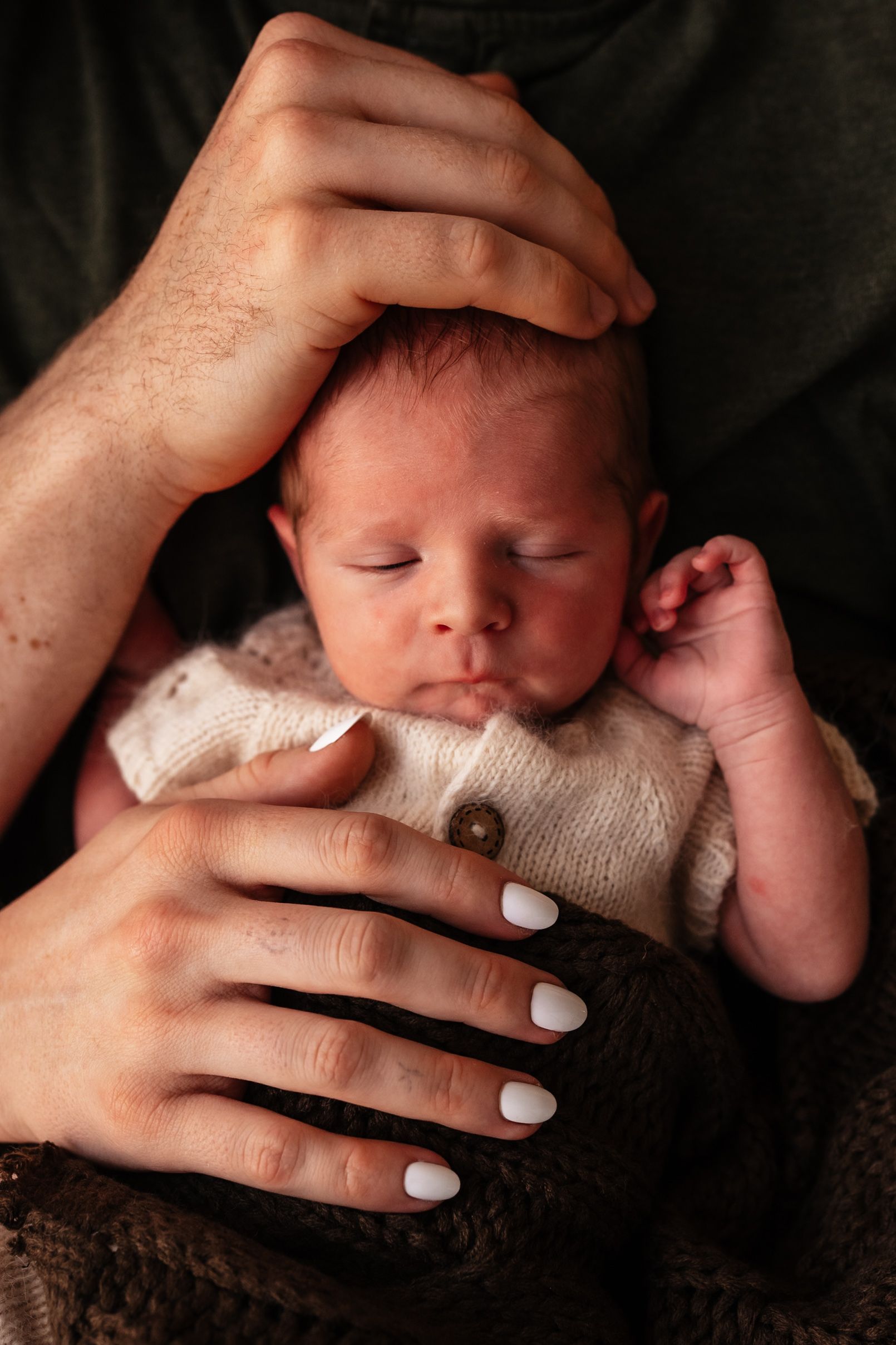 Newborn baby sleeping peacefully cradled in parents hands, with white manicured nails visible.