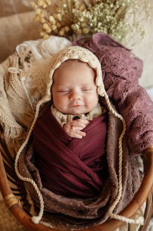 Sleeping newborn baby wrapped in burgundy swaddle wearing cream crochet bonnet nestled in wicker basket with floral decor.