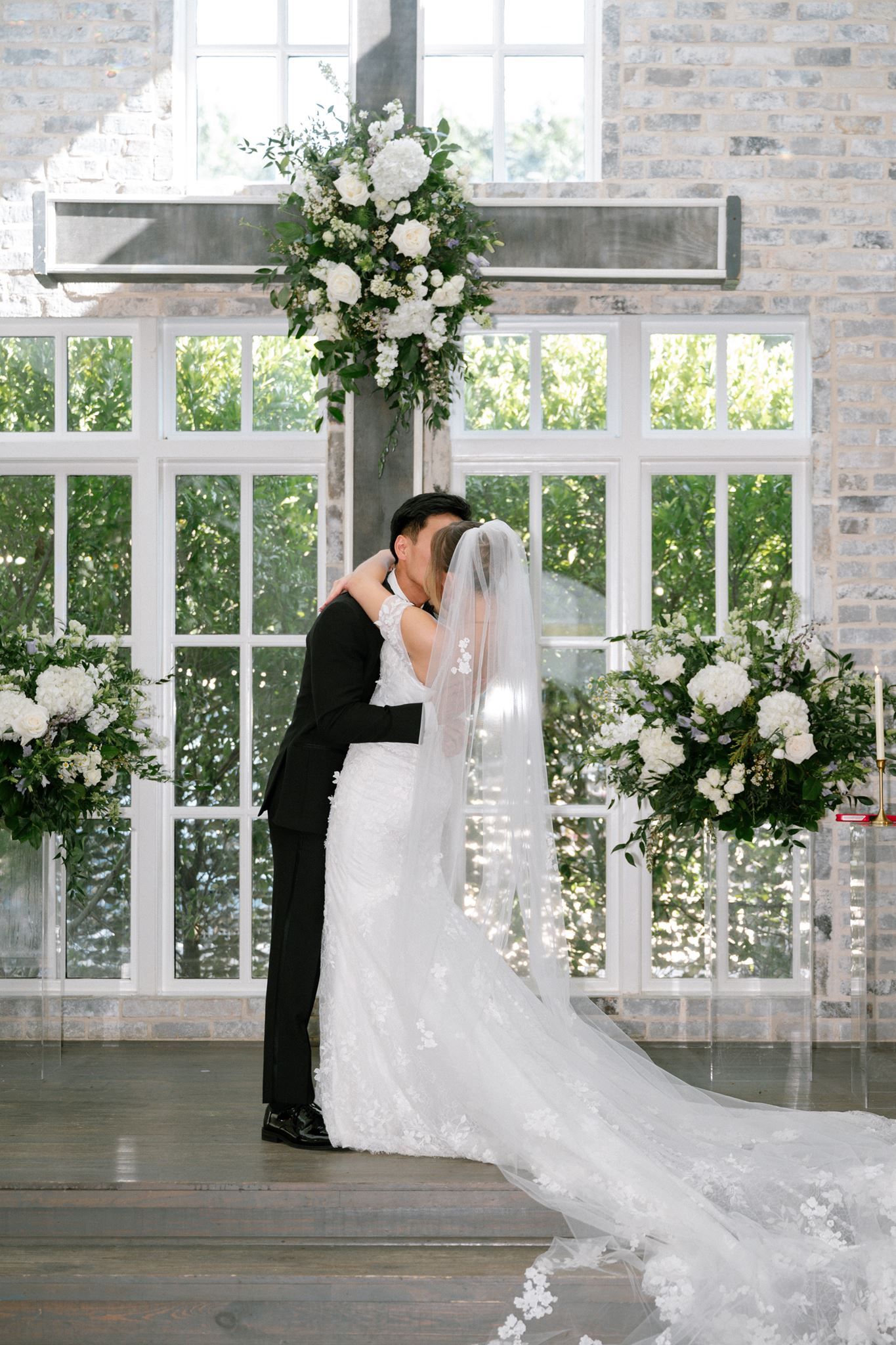 Bride and groom share first kiss at elegant chapel altar adorned with white floral arrangements and a large cross.