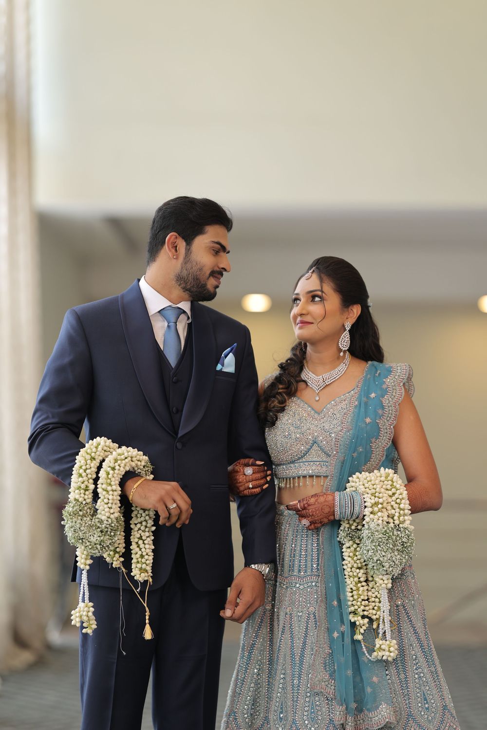Indian couple in wedding attire, groom in navy suit, bride in blue lehenga with floral garlands, sharing a romantic moment.