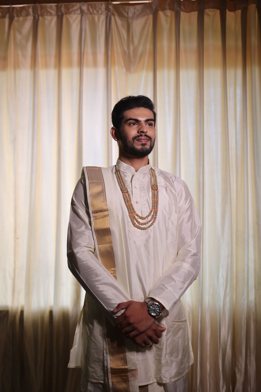 A man in traditional white and gold Indian attire with beaded necklace and watch stands before sheer curtains.