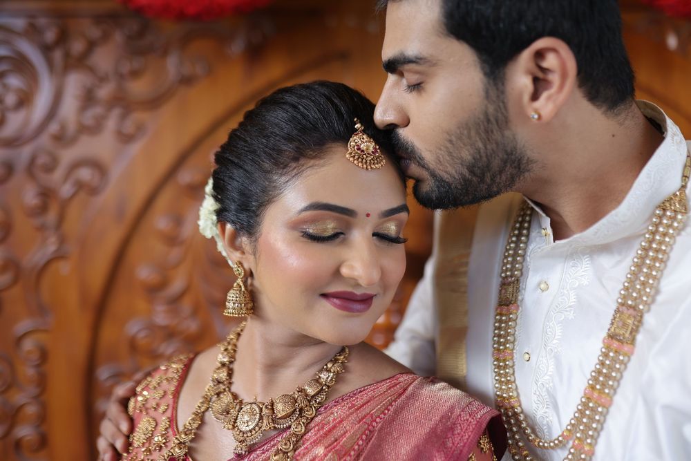 Indian bride and groom in traditional wedding attire, groom kissing bride's forehead, adorned with gold jewelry and garlands.