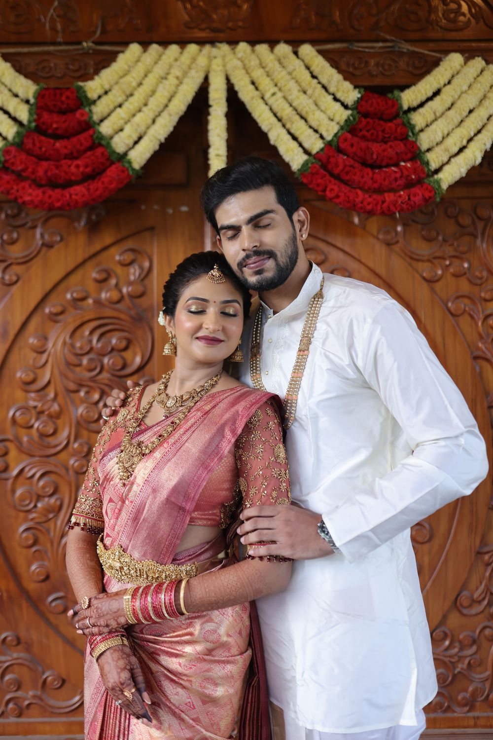 Indian couple in traditional wedding attire, bride in pink silk saree with gold jewelry, groom in white kurta, floral backdrop.
