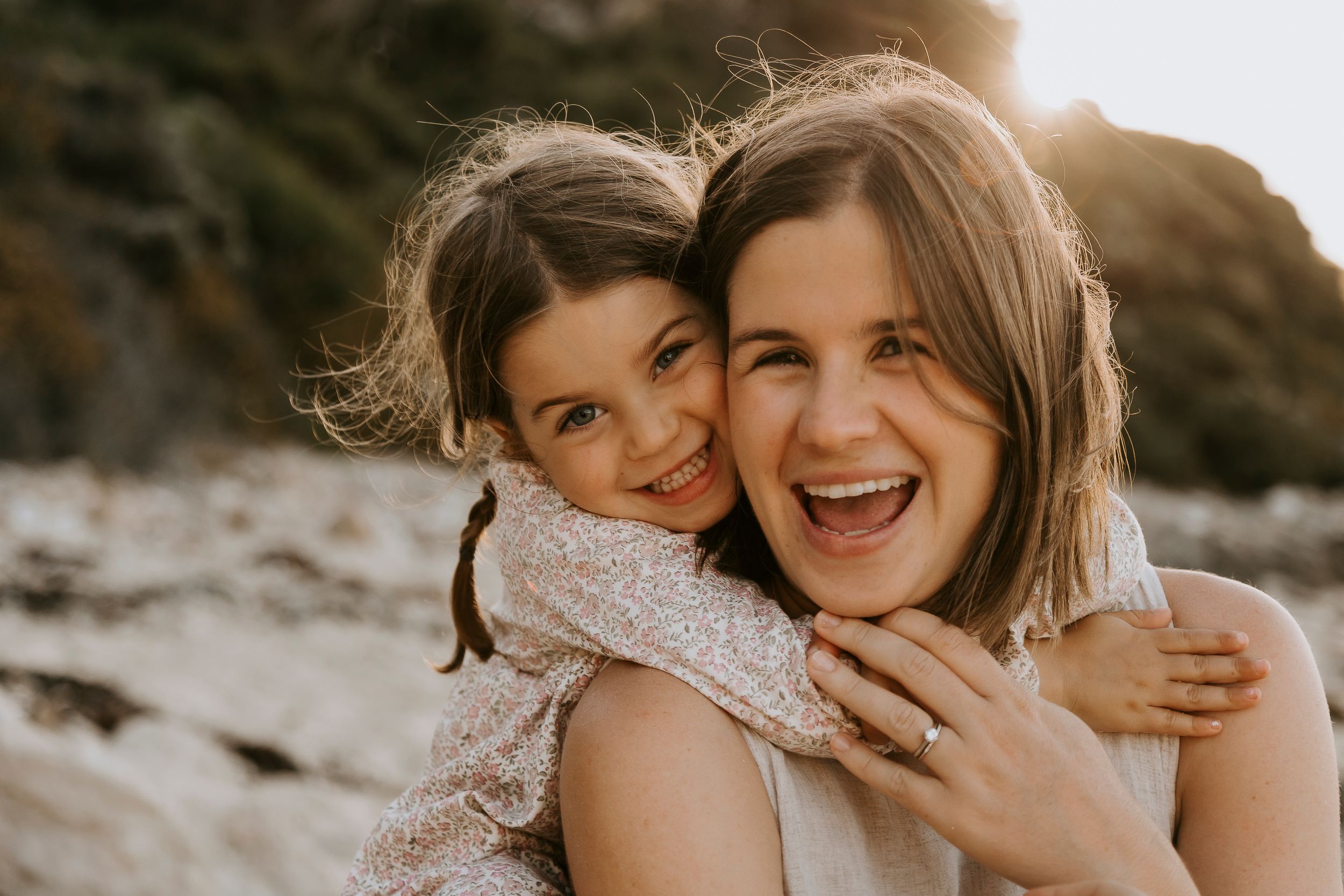 A joyful young girl hugs a laughing woman from behind on a sunny beach, both smiling brightly.