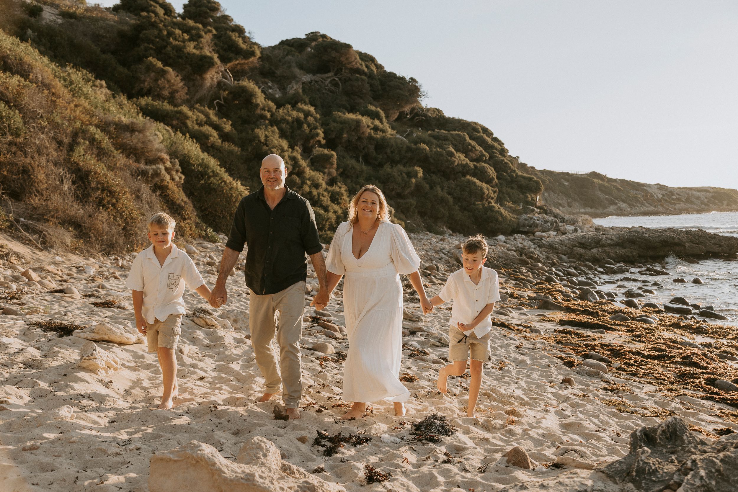 Family of four walking on a sandy beach at golden hour, with rocky cliffs and green hills in the background.