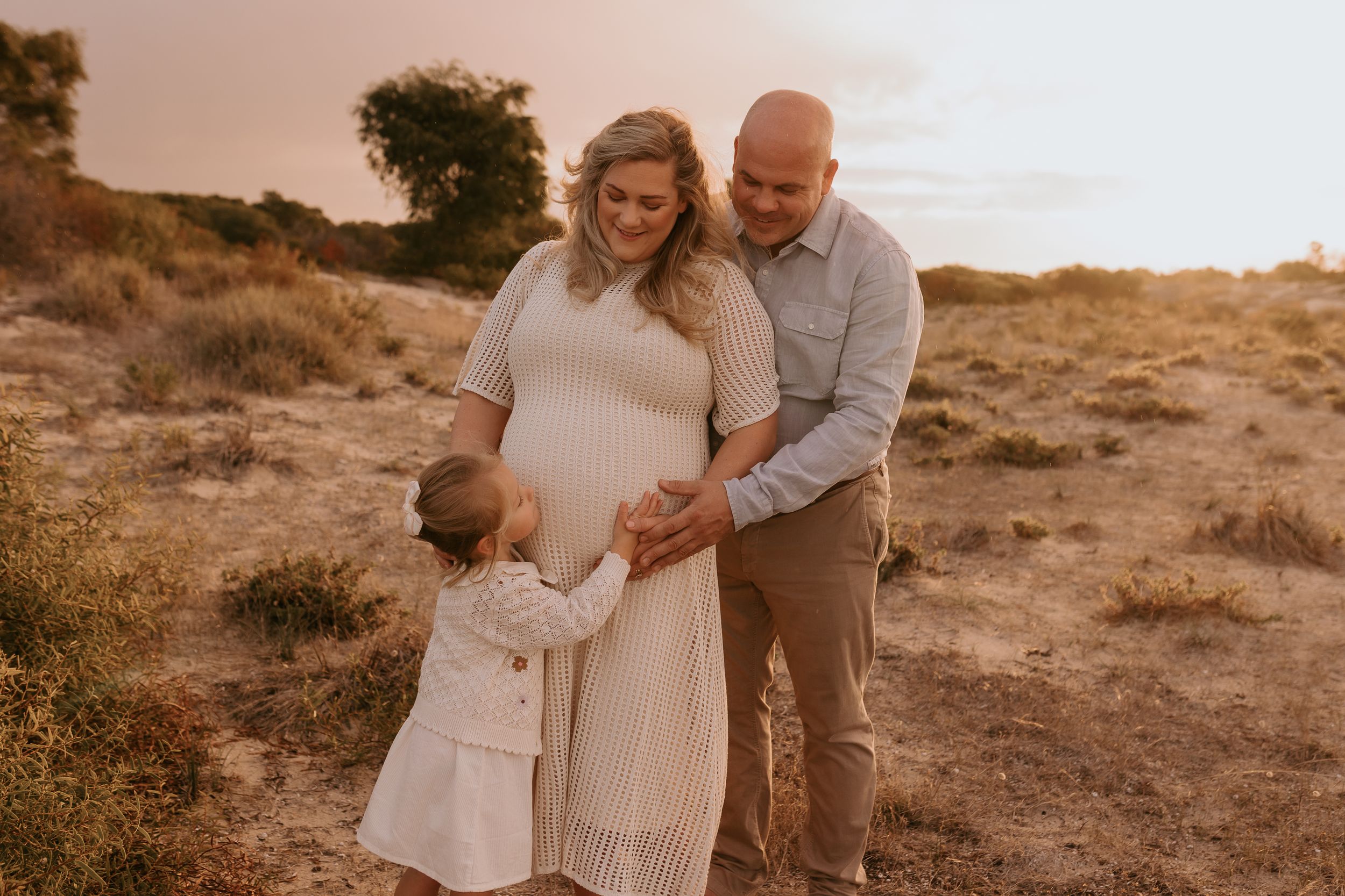 Pregnant woman in white dress poses with husband and toddler in golden field during maternity photo session.