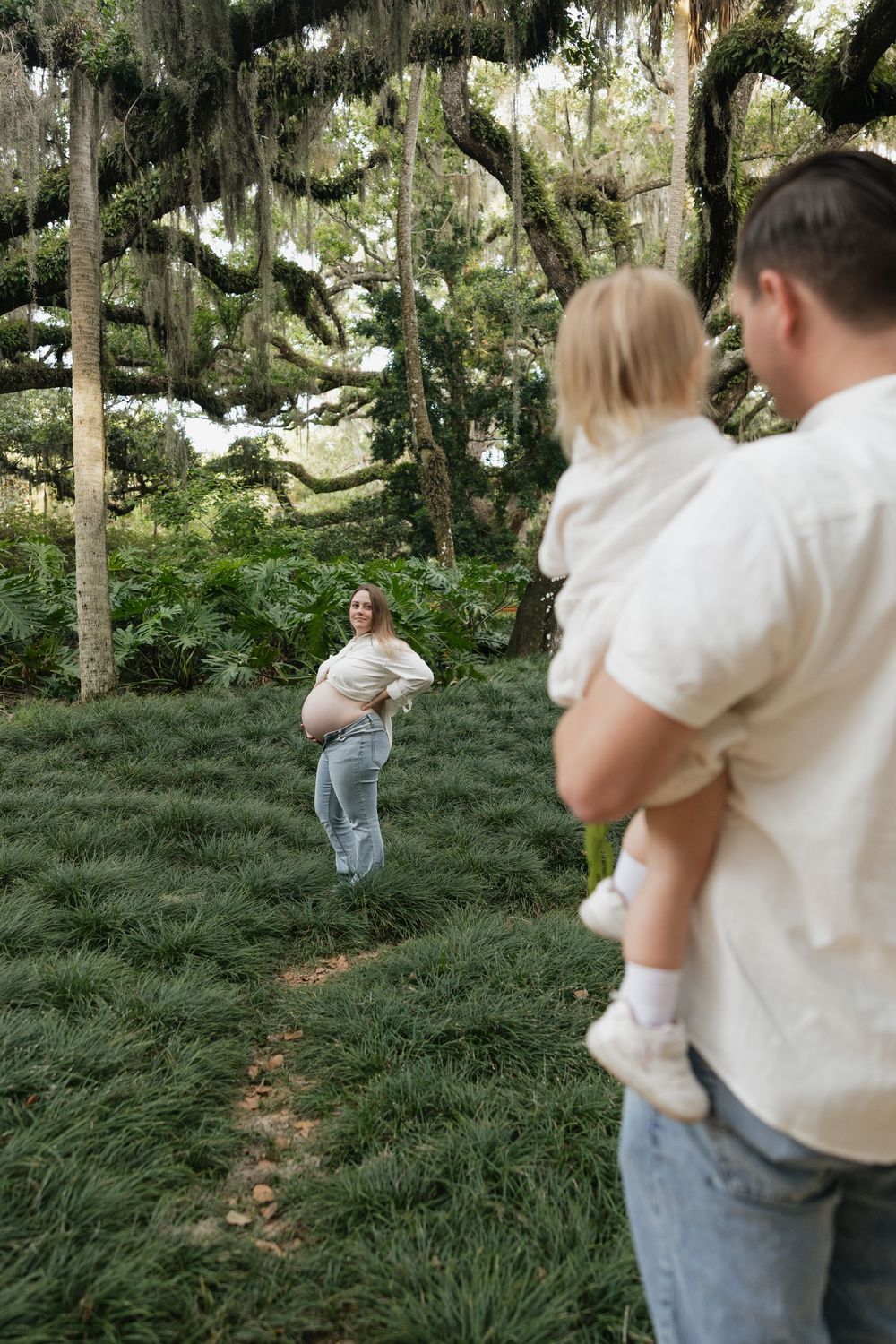 Pregnant woman stands on garden path as man holds toddler under moss-draped oak trees in lush green park.