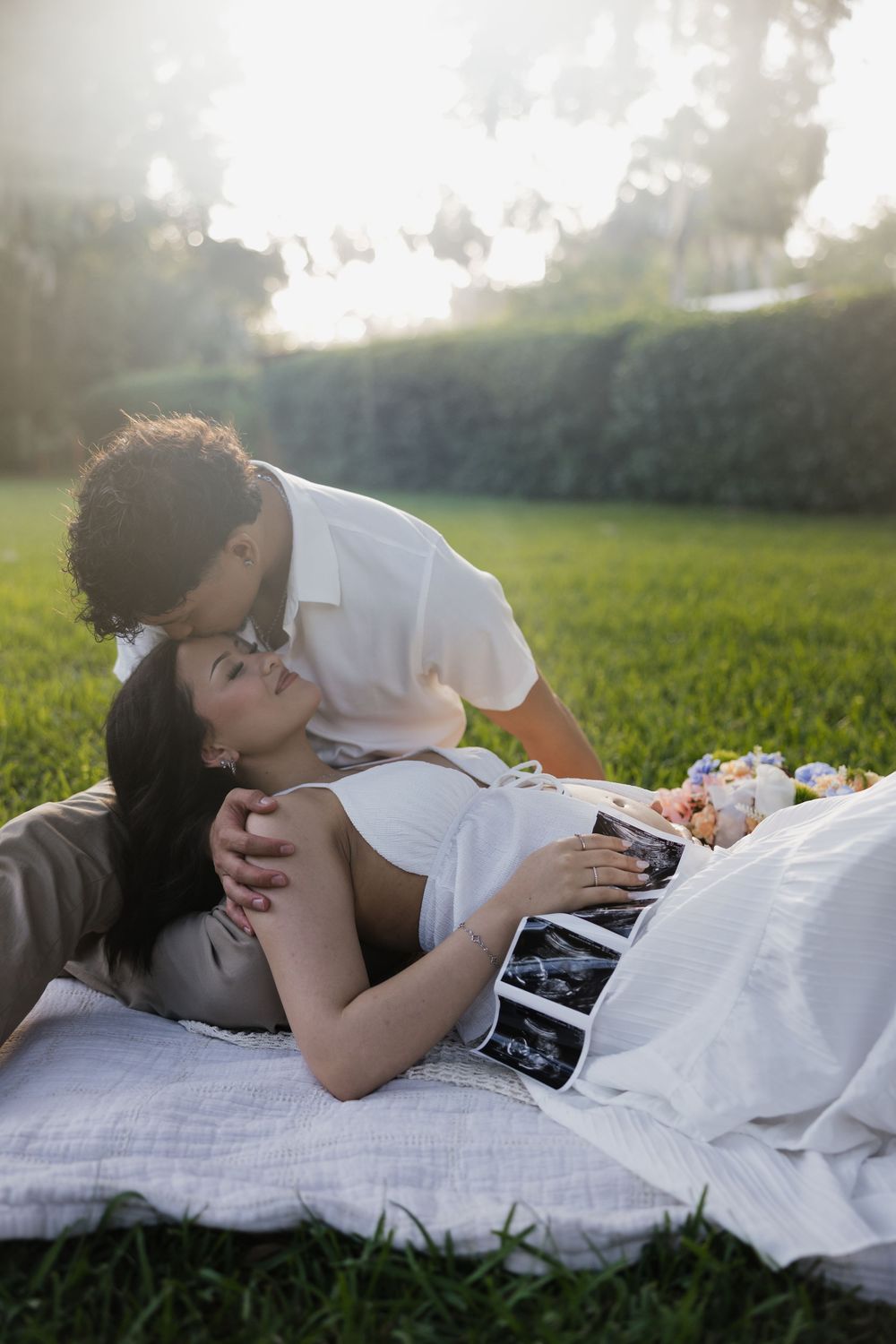 Couple lying on blanket in sunny park, man kissing pregnant woman holding ultrasound photos outdoors.