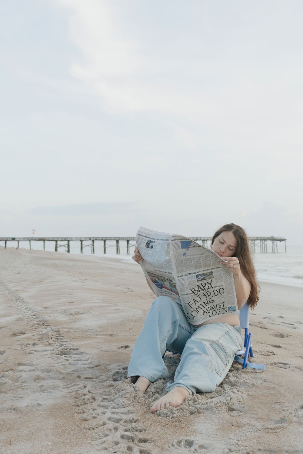 Young woman sitting on sandy beach reading newspaper near pier on overcast day wearing blue jeans.
