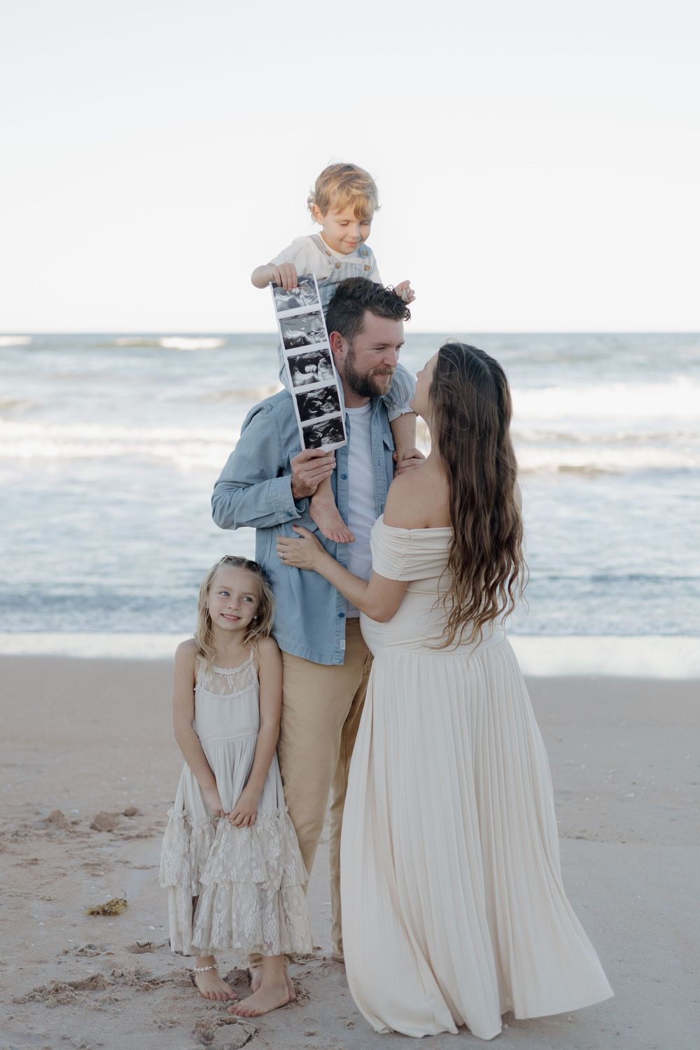 Family of four on beach, parents holding ultrasound photos, toddler on dad's shoulders, girl in lace dress beside them.