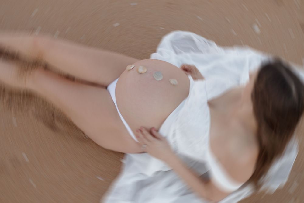 Pregnant woman lying on sand beach in white bikini with decorative stones on baby bump, aerial view.