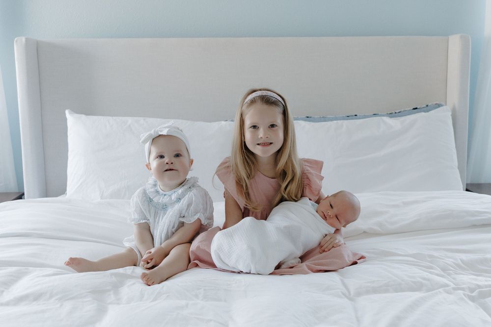 Three young sisters pose on a white bed, with the oldest holding a swaddled newborn baby.
