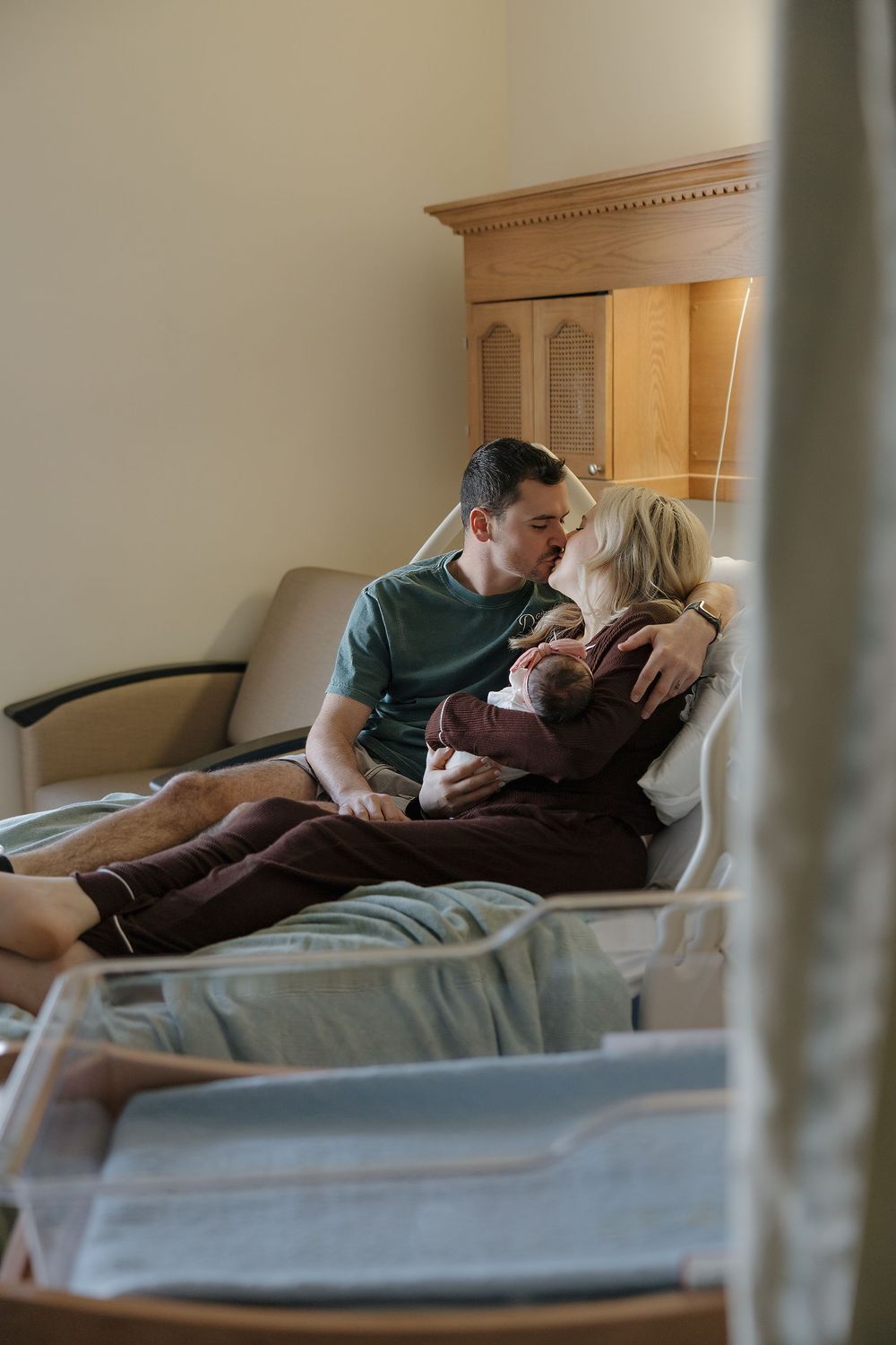 New parents share a tender kiss in a hospital room while holding their newborn baby together on the bed.