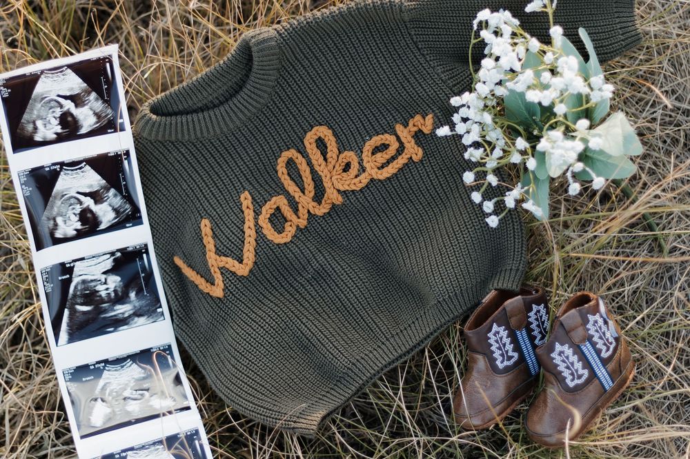 Olive green knit baby sweater with name Walker, ultrasound photos, tiny boots, and white flowers on dried grass.