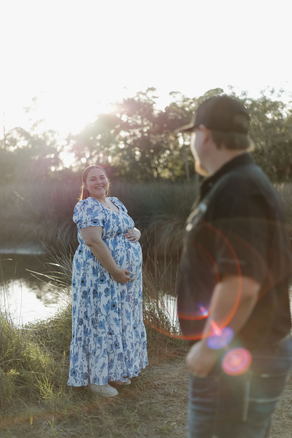 Pregnant woman in blue floral dress smiles outdoors near a pond at golden hour with a man in foreground.