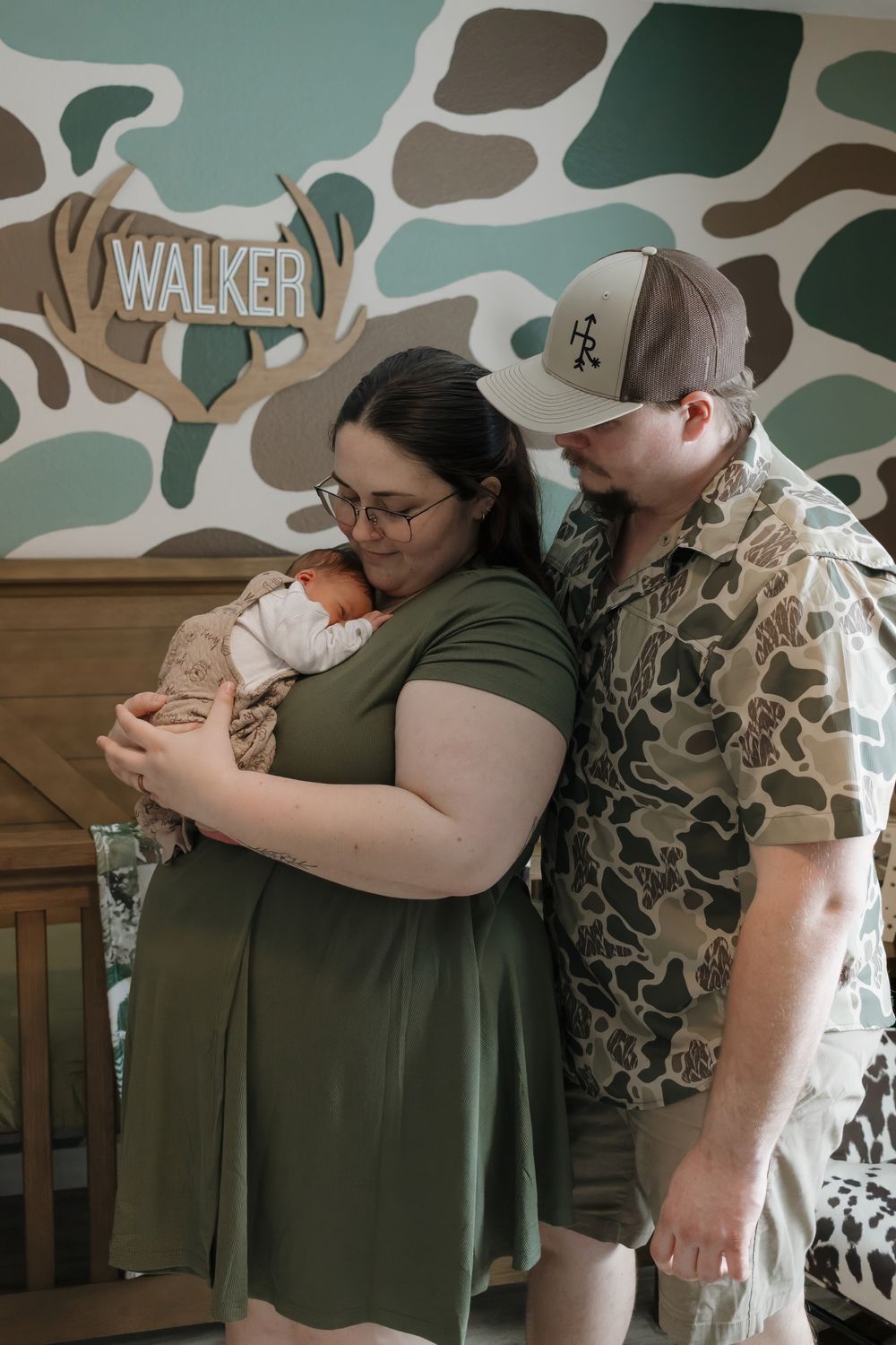 New parents in camo outfits tenderly hold their newborn baby in a hunting-themed nursery with Walker name sign.