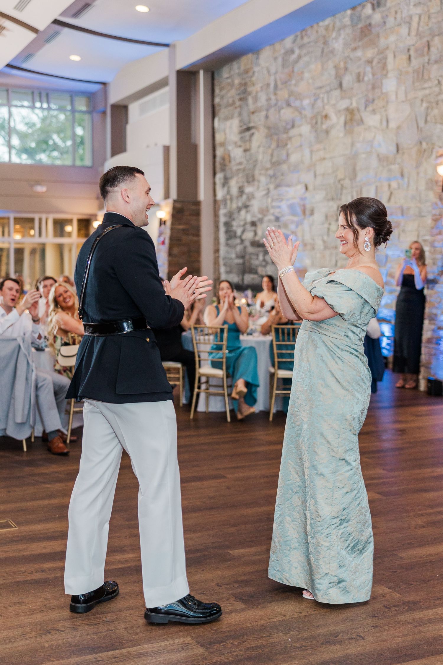 A military groom and his mother share a joyful dance at an elegant wedding reception with stone wall backdrop.
