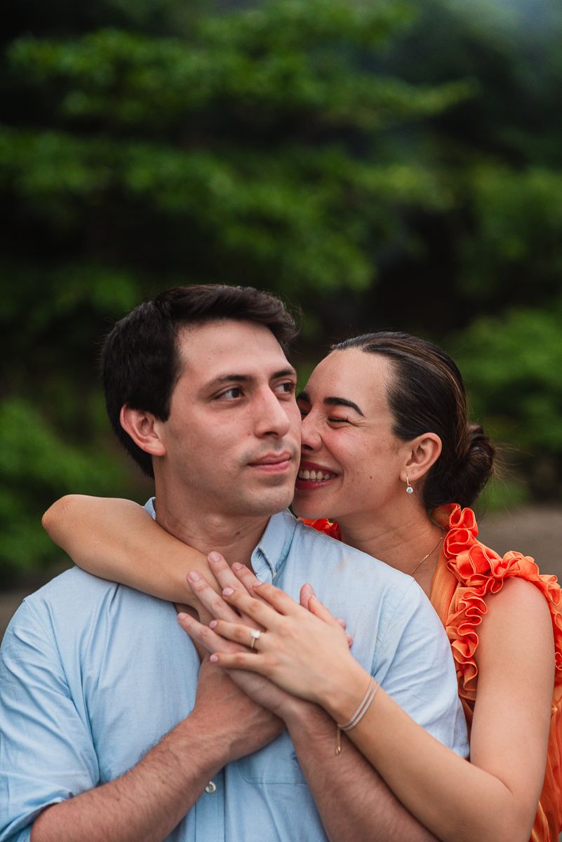 A smiling woman in an orange dress embraces a man in a light blue shirt outdoors with green trees behind them.