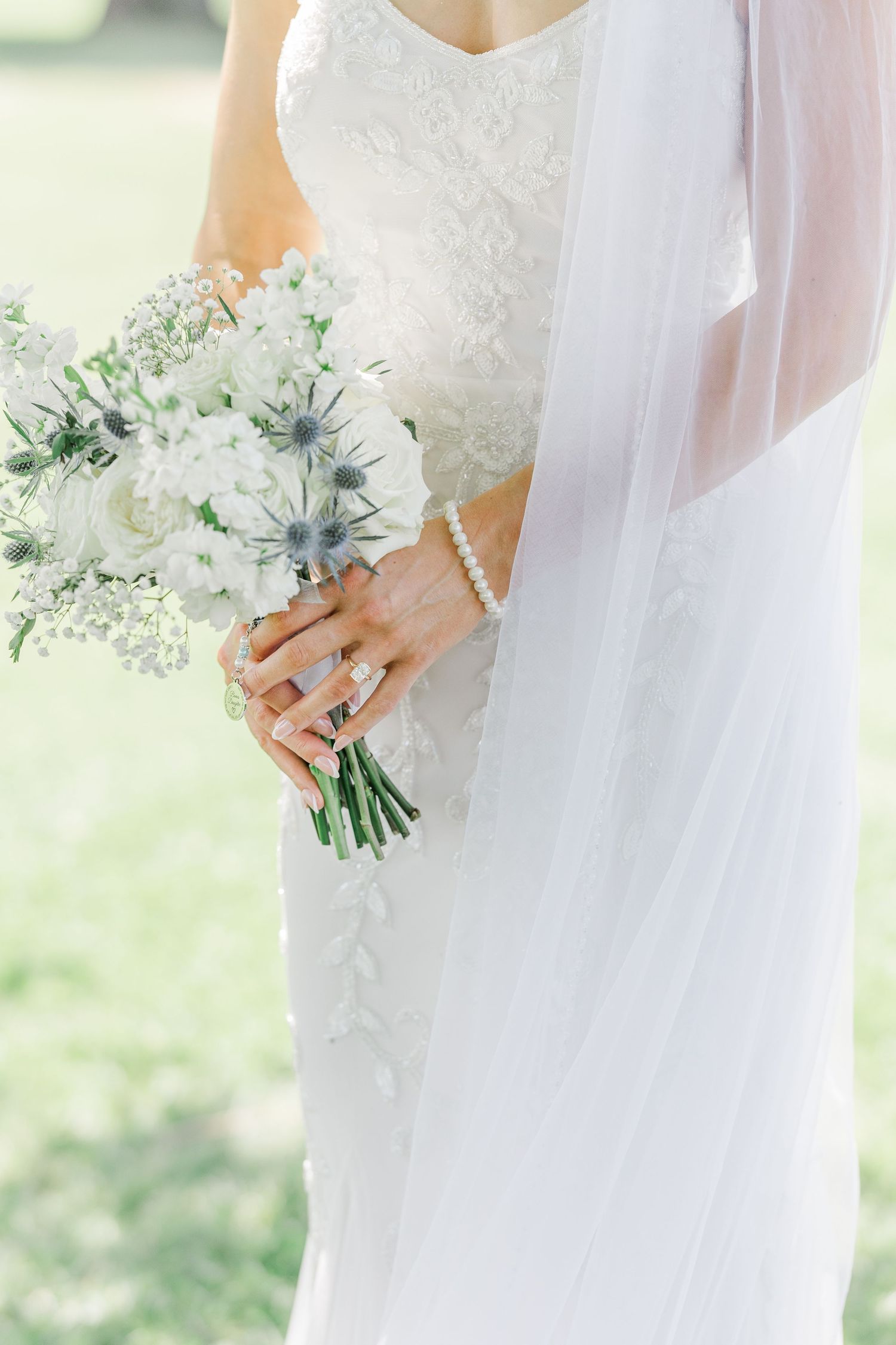 Bride in lace wedding gown holding white and blue thistle bouquet with pearl bracelet and diamond ring.