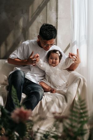 Father playfully scaring smiling baby girl in white dress while sitting together on a couch near a sunlit window.父親和身穿白色洋裝、笑容燦爛的女嬰坐在陽光照射的窗邊