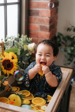 Joyful baby laughing in a wooden tub with rubber duck, orange slices, sunflowers, and soap bubbles.快樂的寶寶在木桶裡咯咯地笑著，桶子裡有橡皮鴨、橘子片、向日葵和肥皂泡。