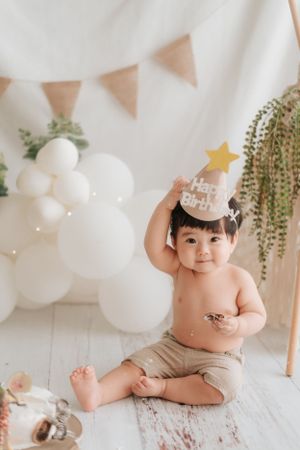 Baby wearing party hat sits on wooden floor with white balloon decorations.戴著派對帽的嬰兒坐在木地板上，周圍裝飾著白色氣球。