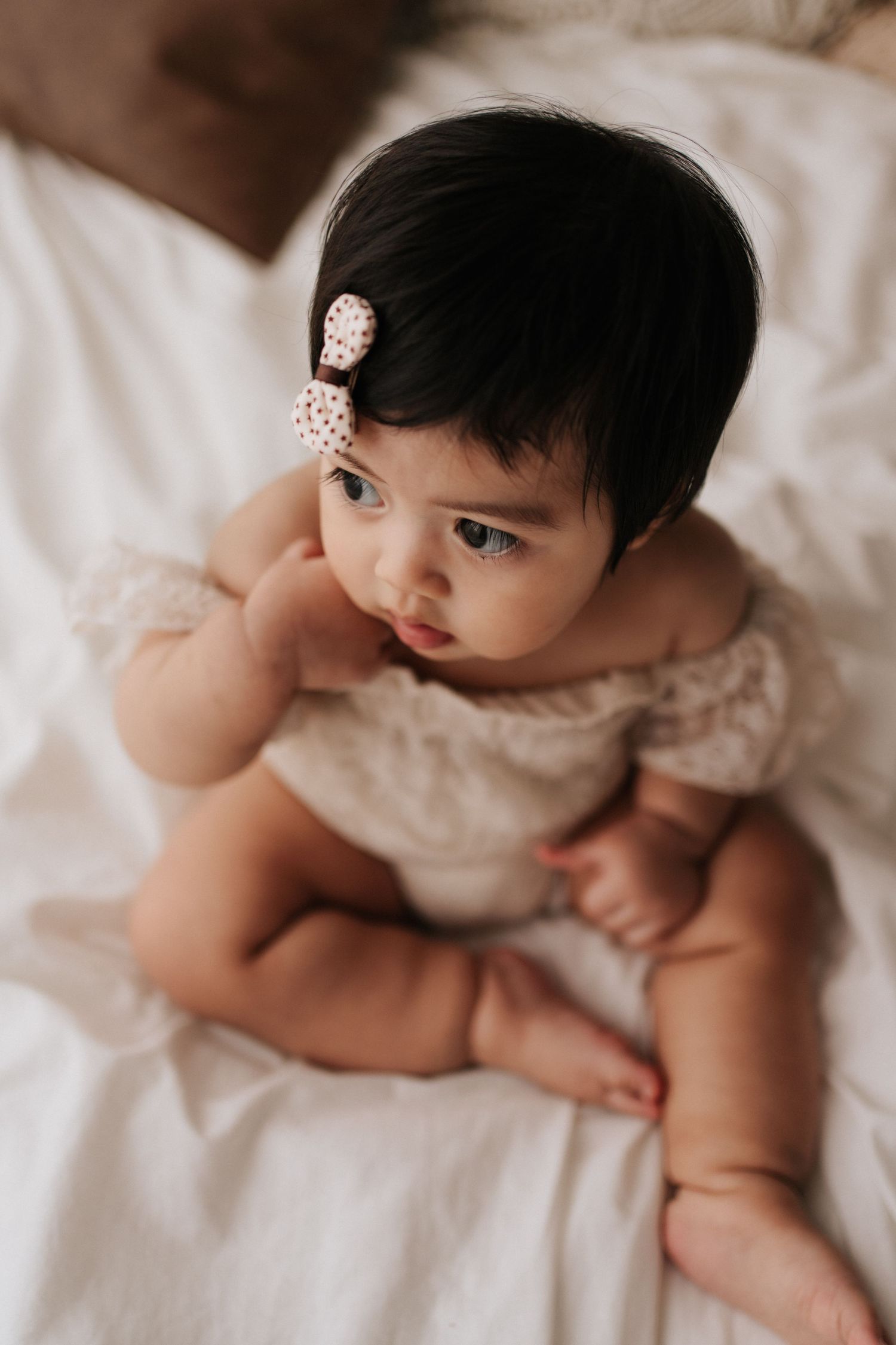 Baby in cream dress with pearl hair clip sitting on white bedding.身穿奶油色洋裝、頭戴珍珠髮夾的嬰兒坐在白色床單上。