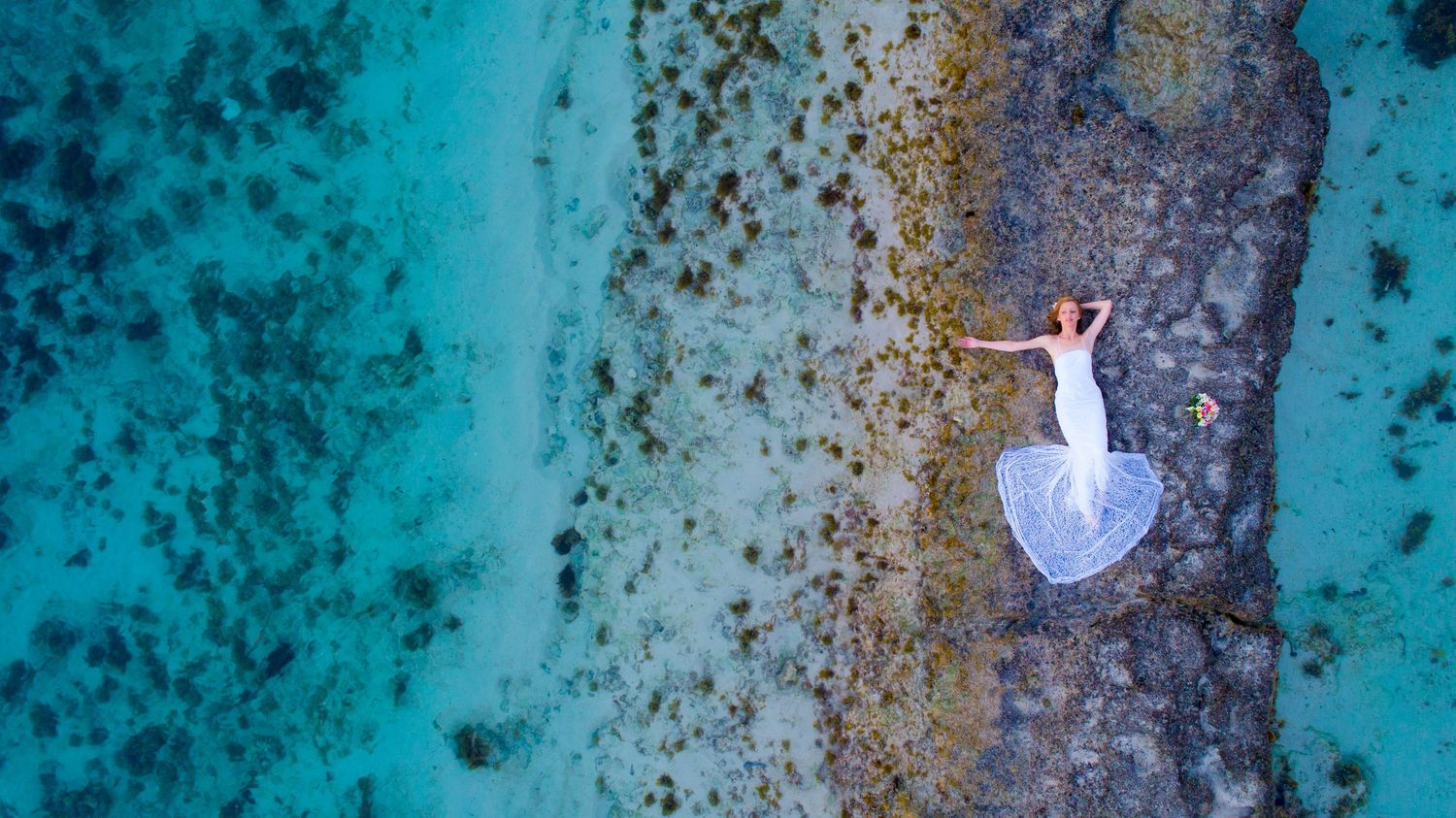 Aerial view of bride in white gown lying on a coral reef surrounded by turquoise lagoon, Mauritius wedding photographer Luminair Studio