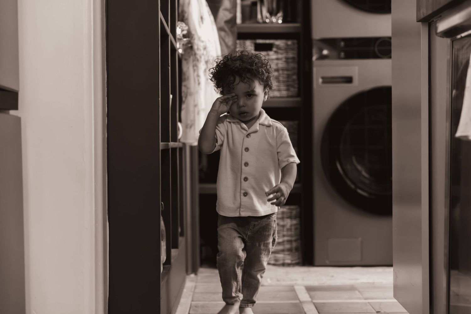 Black and white photo of a young child walking through a laundry room hallway with a washing machine in background.