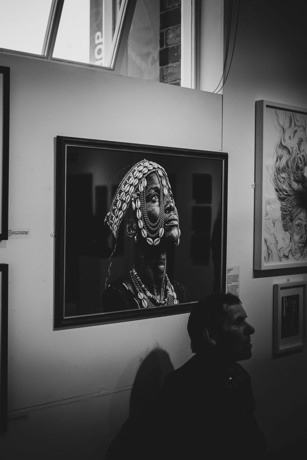Black and white photo of a person in an art gallery viewing a framed portrait of a tribal woman with traditional jewelry.