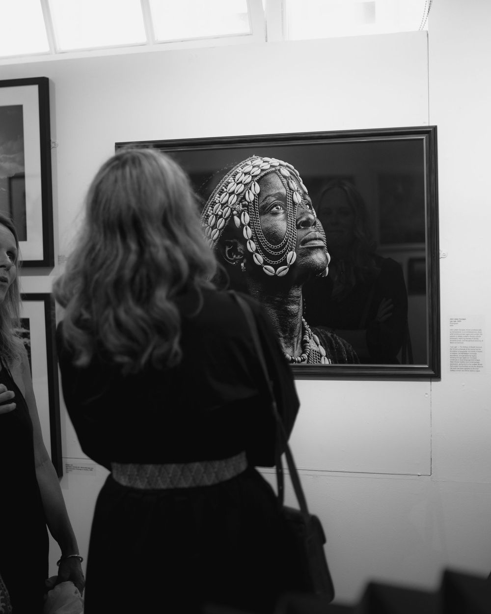 Woman with long hair views a framed black and white portrait of a tribal elder at an art gallery exhibition.